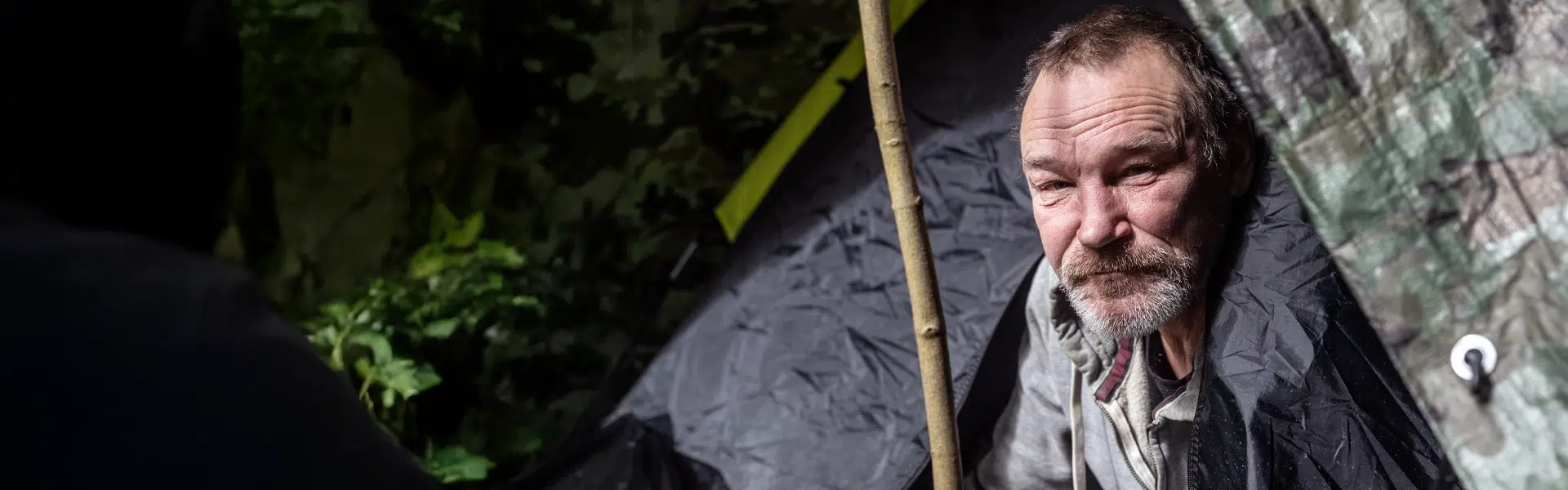 A man in the opening of a make shift tent in a wooded area.
