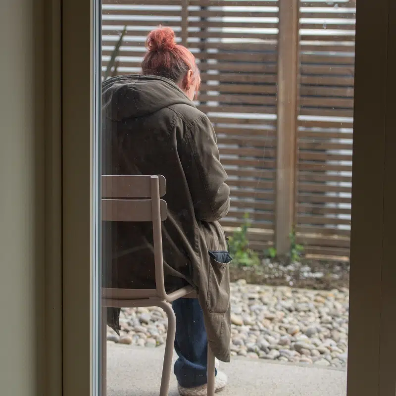A view from inside looking at a woman wearing a coat who is sitting outside with her back to the viewer