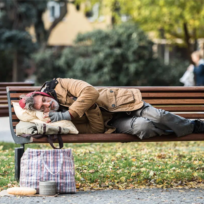 A man sleeps on a park bench in the day time. He is using bags as a pillow and has other bags with him.