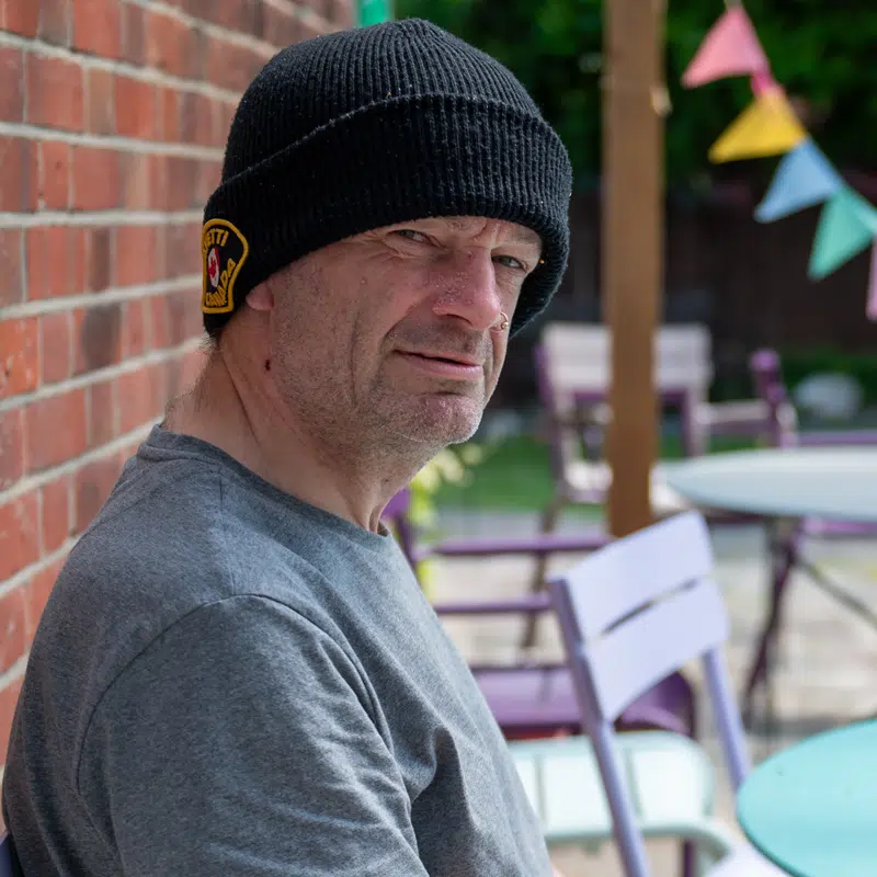 A middle aged man wearing a woolie hat sits in a garden